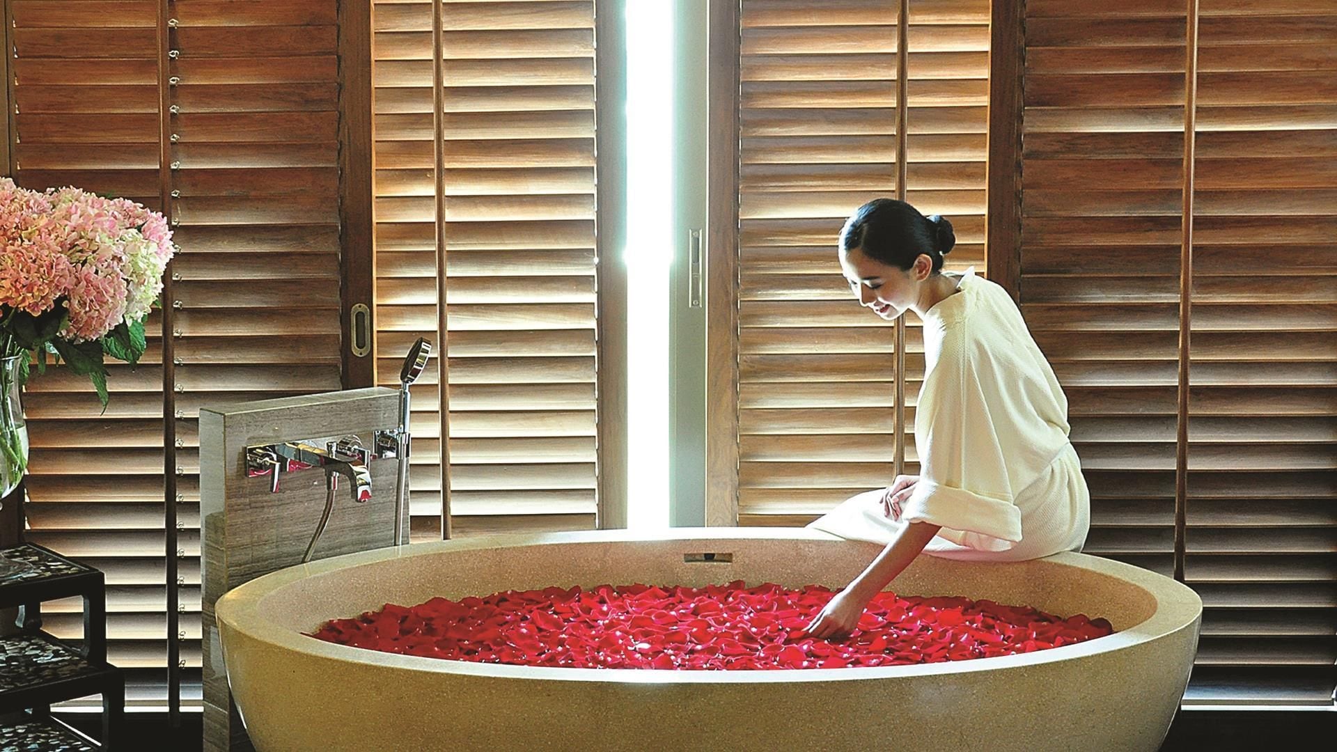 Une personne assise dans une grande baignoire remplie de pétales de roses rouges, avec des stores en bois en arrière-plan.