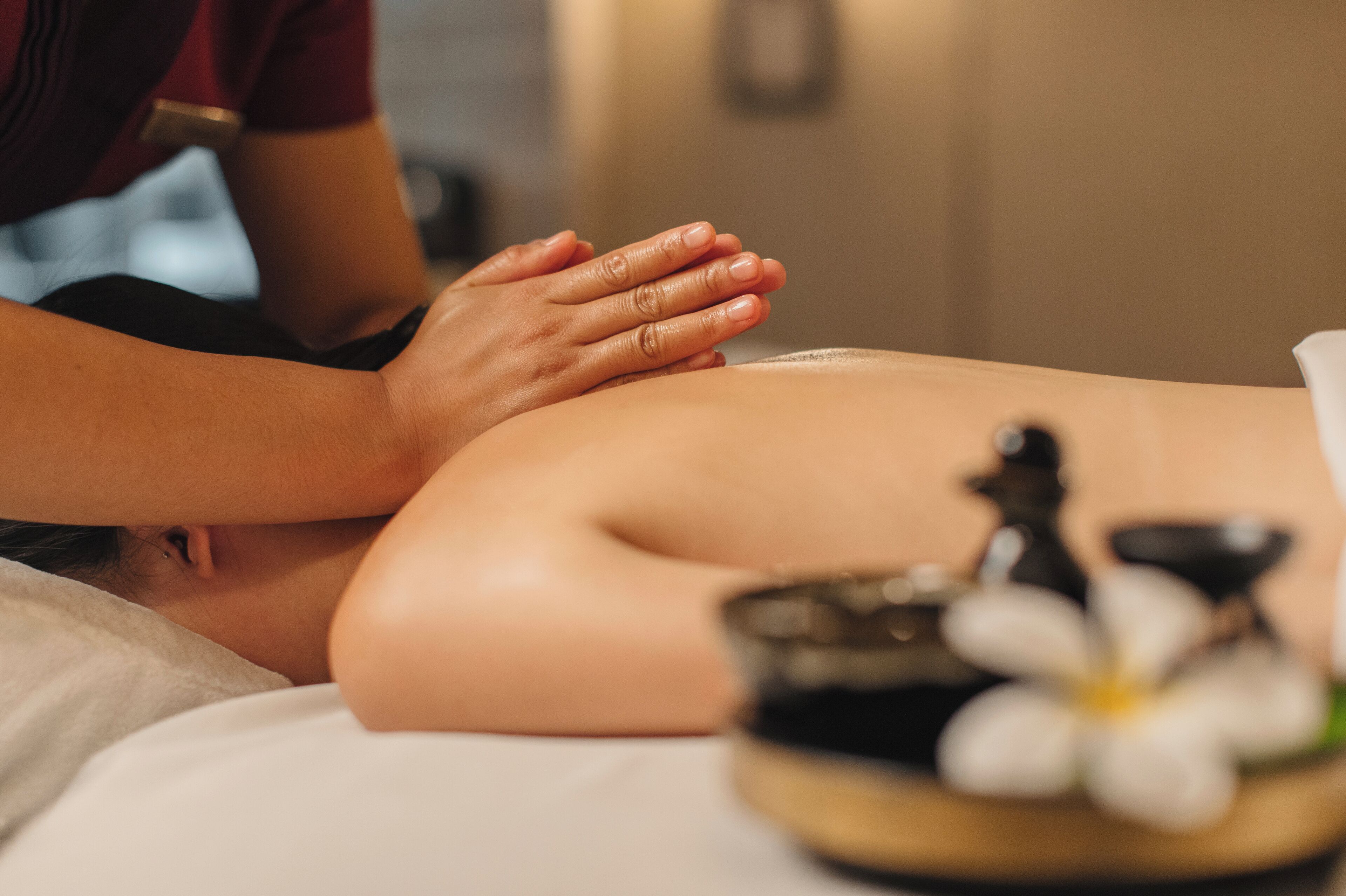 A massage therapy session with a person lying face down, a therapist's hands on their back, and a small bowl with a flower on a table beside them.