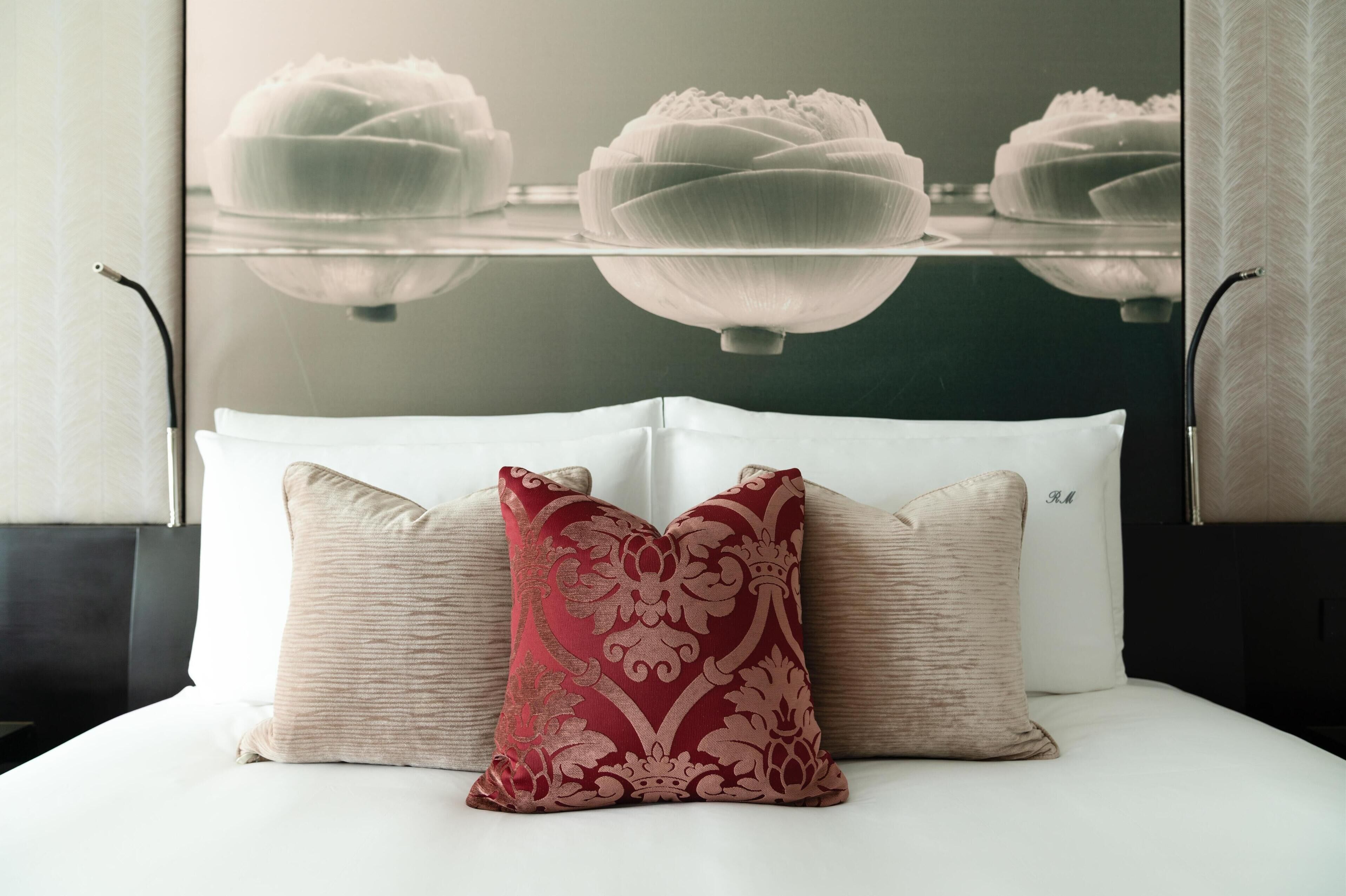 A bed with white linens and two pillows, one red and one beige, in a modern room with a glass partition and a reflection of a lotus flower.