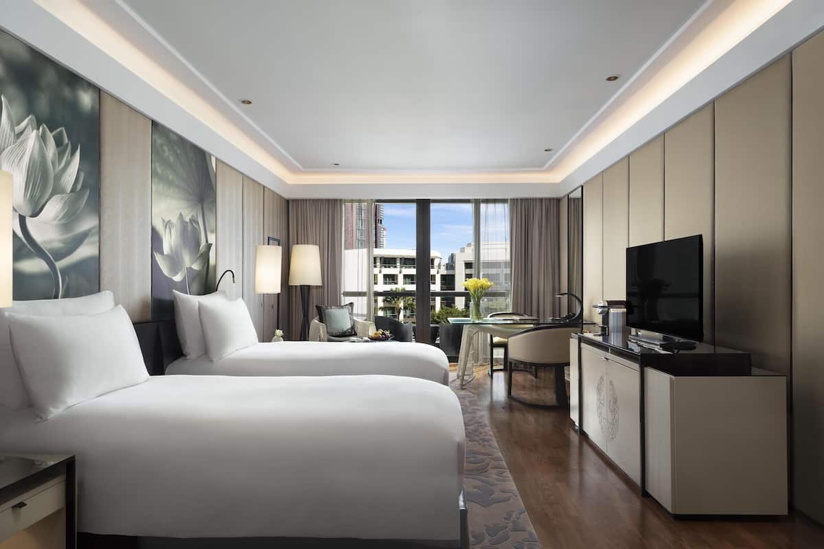 Ein Hotelzimmer mit zwei Betten, einem Schreibtisch mit Fernseher und einem großen Fenster mit Blick auf die Stadt.
