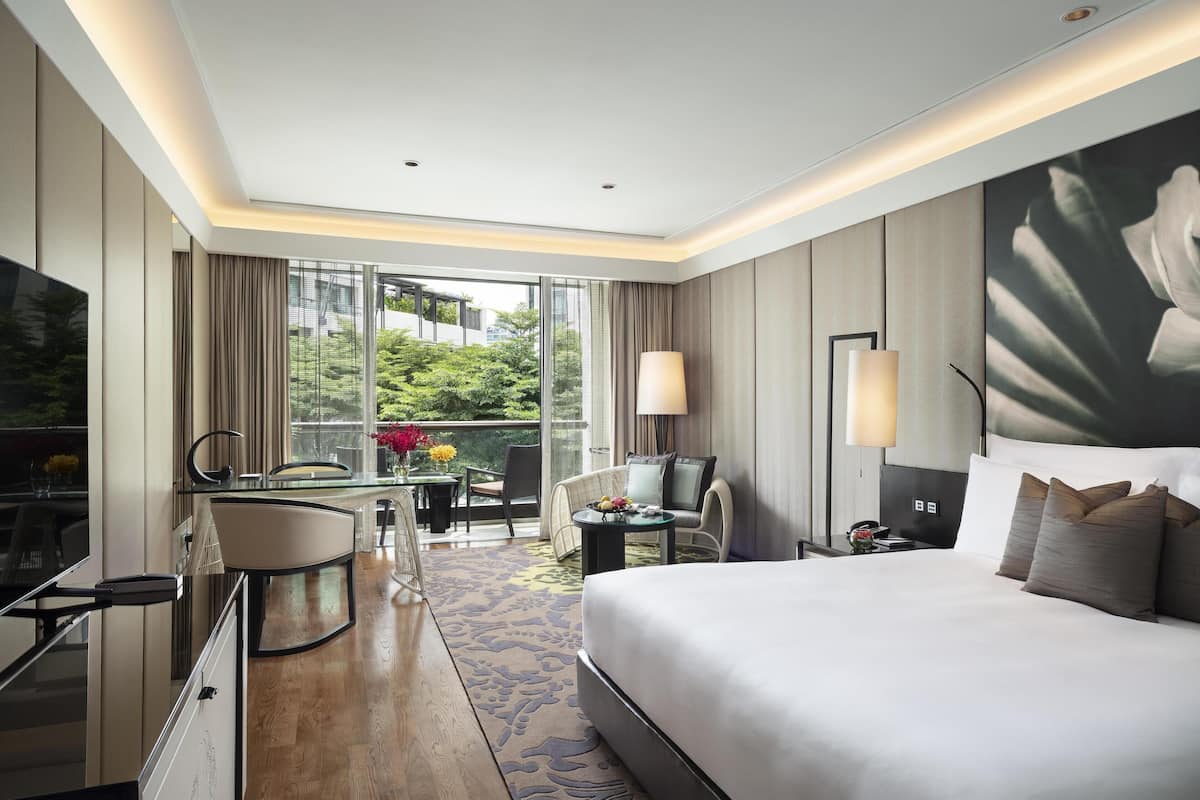 Ein Hotelzimmer mit einem großen Bett, einem Schreibtisch mit Stuhl, einem Fernseher und einem Balkon mit Blick auf Grünflächen.