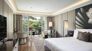 A hotel room with a large bed, a desk with a chair, a TV, and a balcony with a view of greenery.