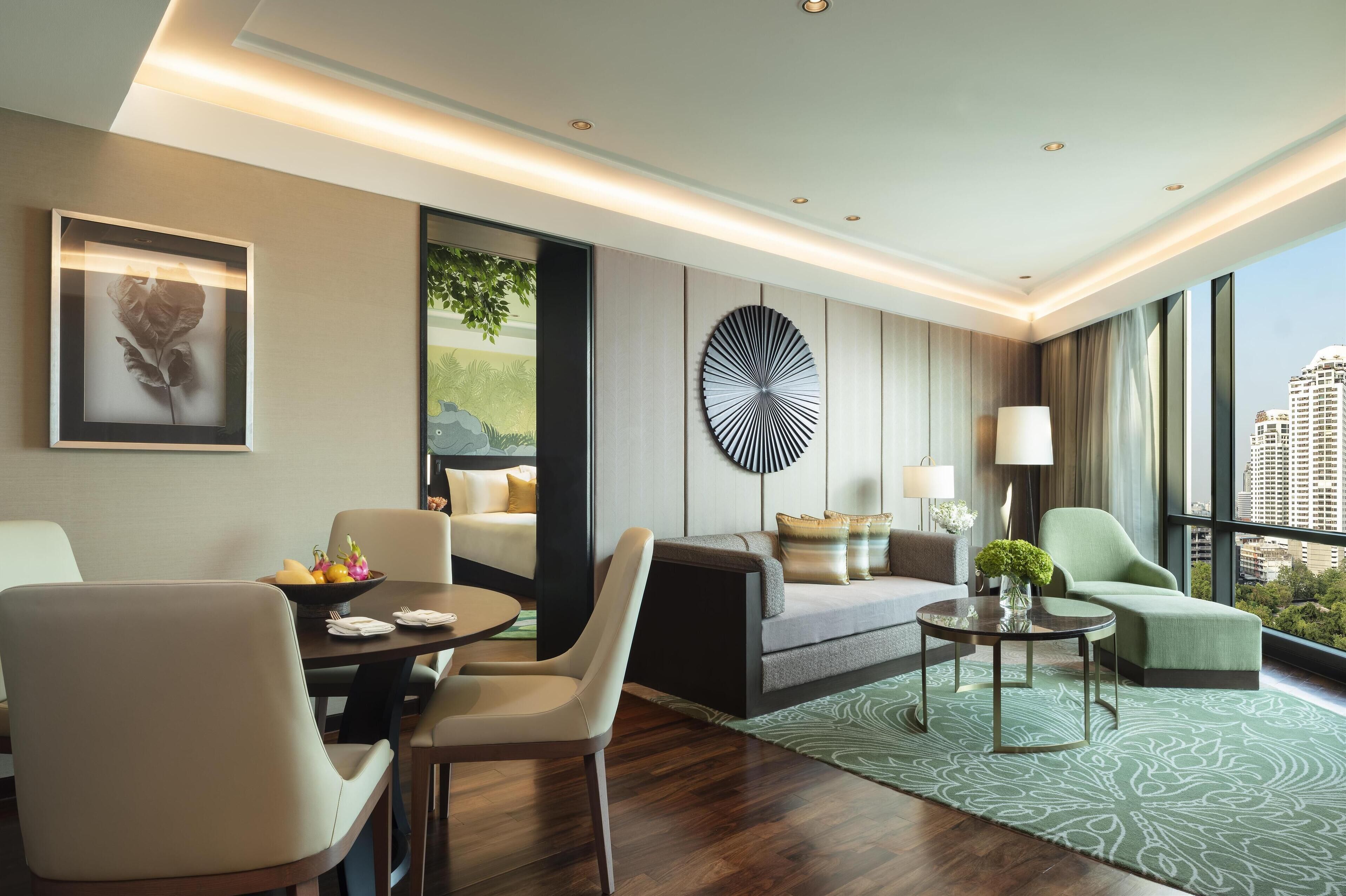 A modern hotel room with a dining area, a sofa, a green armchair, and a view of the city.