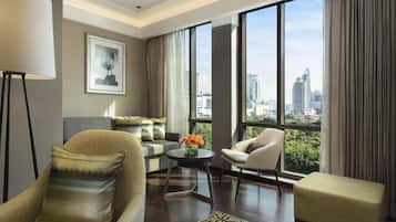 A modern living room with a sofa, armchair, and ottoman, a coffee table with flowers, and large windows offering a city view.