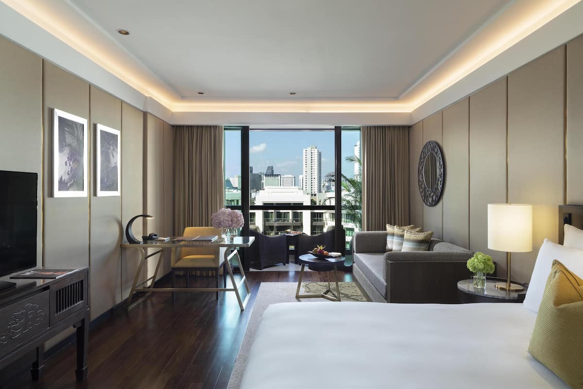 Ein modernes Hotelzimmer mit einem großen Bett, einem Schreibtisch, einer Couch und einem Fernseher. Das Fenster bietet einen Blick auf umliegende Gebäude.