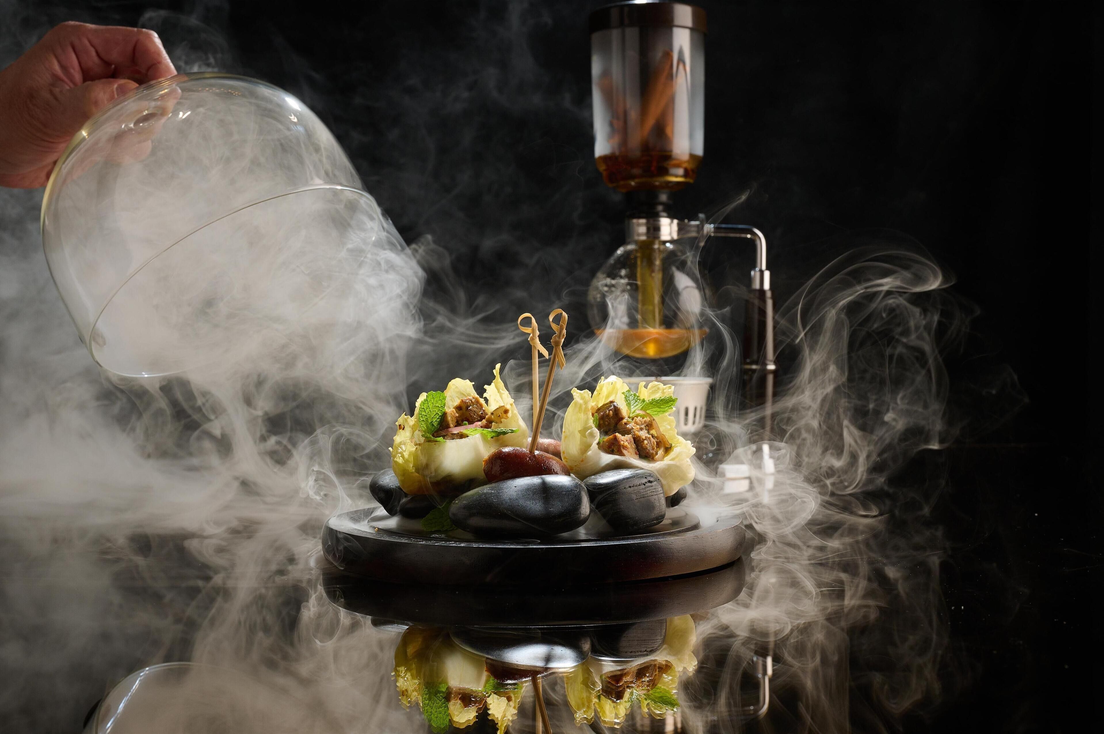 A hand pouring smoke into a smoking device with a bowl of food and rocks.