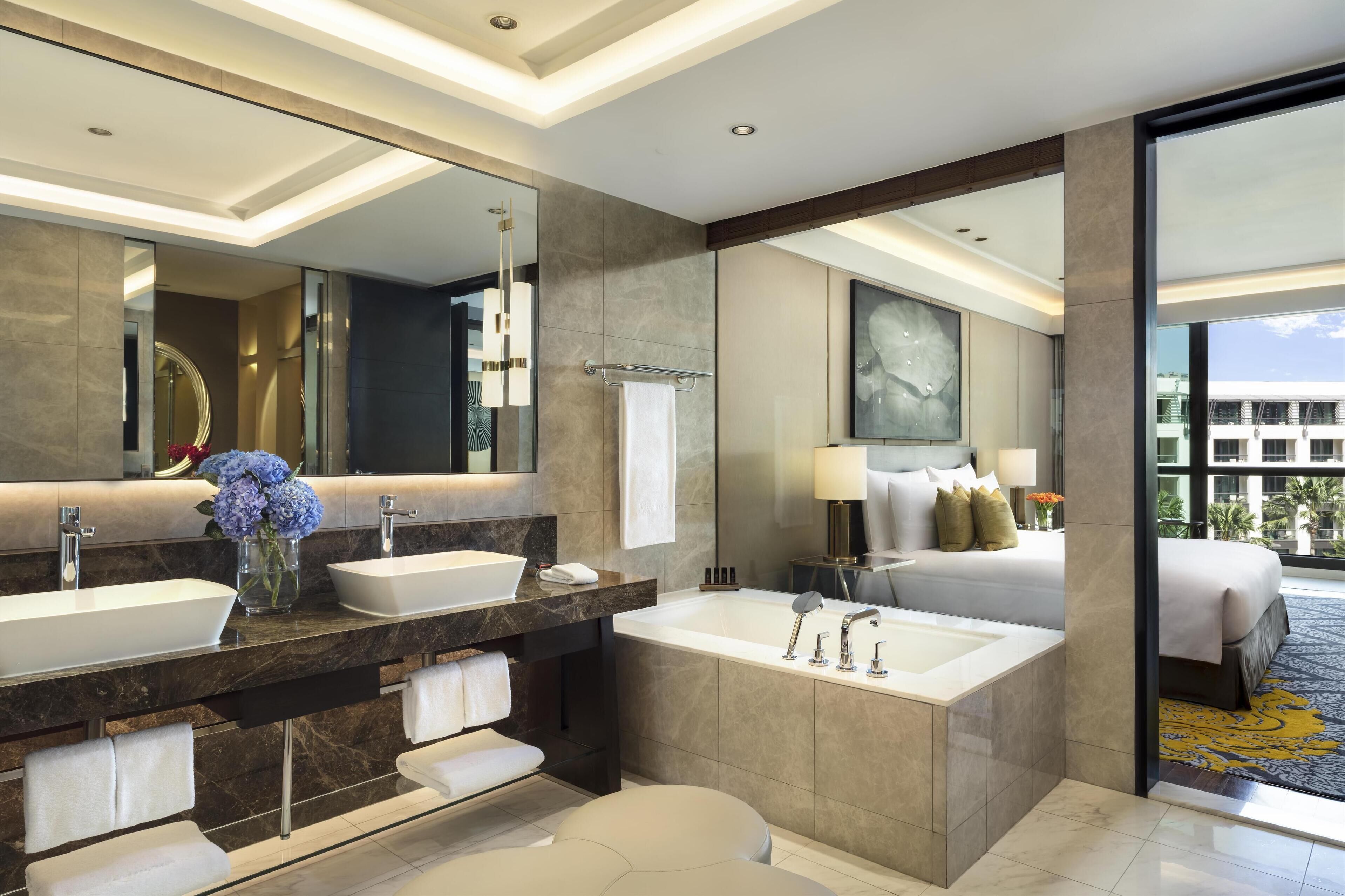 A modern hotel bathroom with dual sinks, a large mirror, a bathtub, and a view of the hotel exterior.