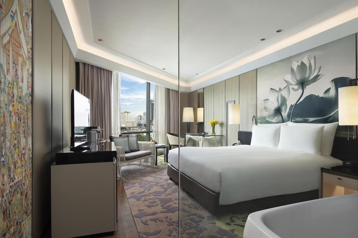 Ein Hotelzimmer mit einem großen Bett, einem Schreibtisch, einem Stuhl, einem kleinen Tisch mit einer Lampe und Blick auf die Stadt.