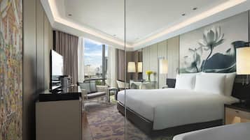 A hotel room with a large bed, a desk, a chair, a small table with a lamp, and a view of the city.