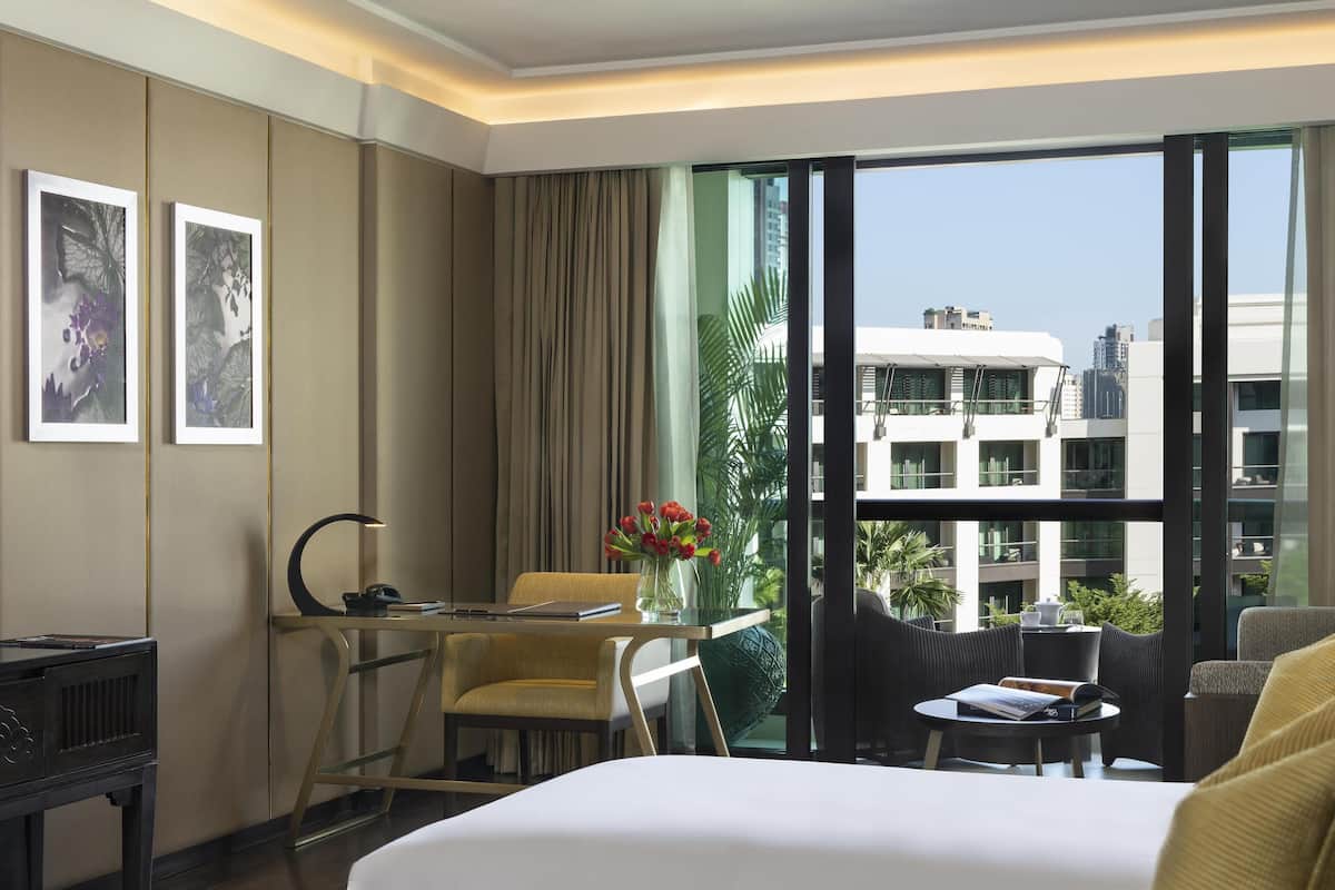 Ein Hotelzimmer mit Bett, Schreibtisch mit Stuhl, einem kleinen Tisch mit Blumenvase und Blick auf die Stadt durch große Fenster.