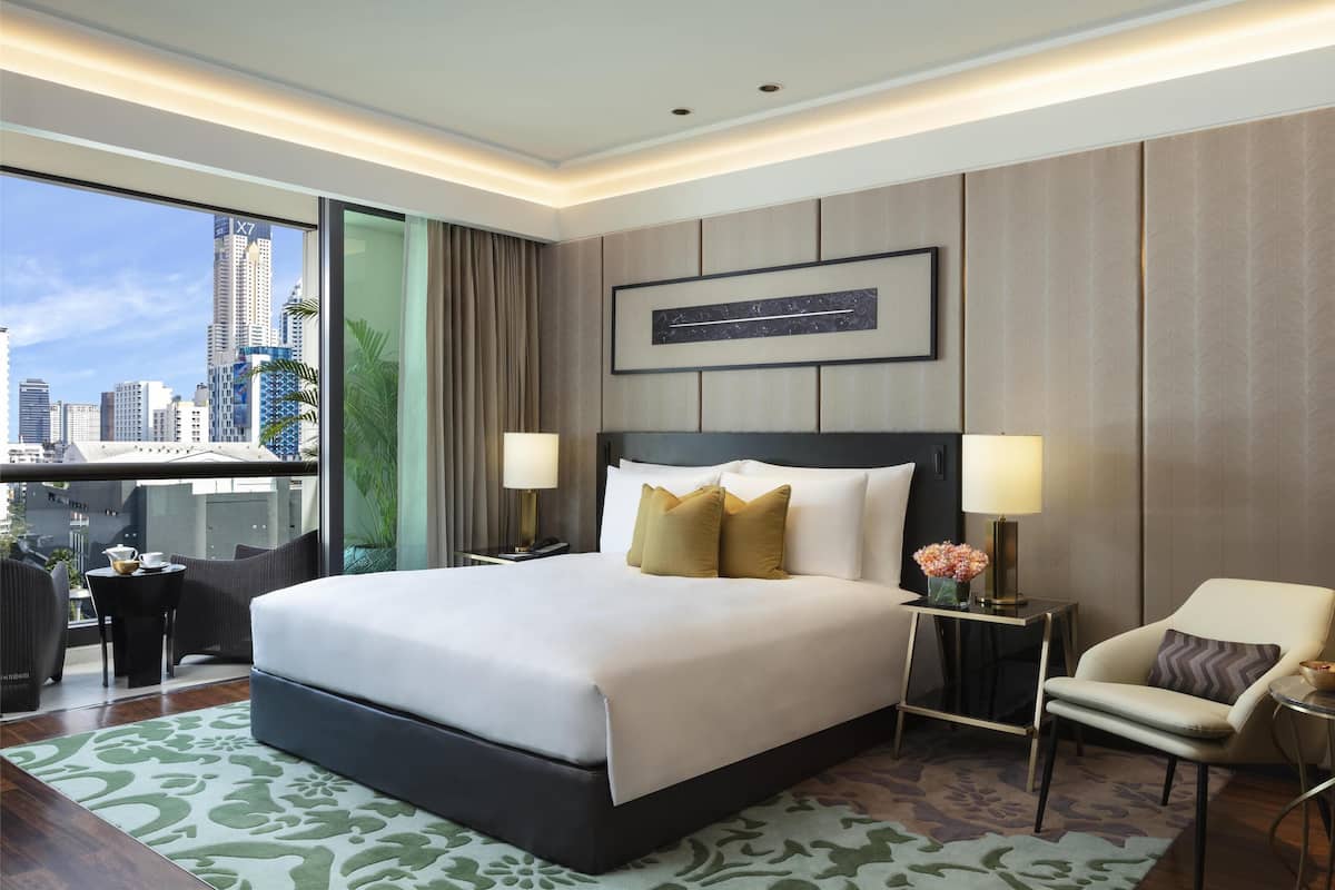 Ein modernes Hotelzimmer mit einem großen Bett, zwei Nachttischen mit Lampen, einem Sessel und Blick auf die Skyline der Stadt durch große Fenster.