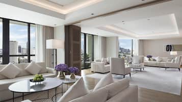 A modern hotel room with a large sofa, armchairs, a coffee table, and a city view through large windows.