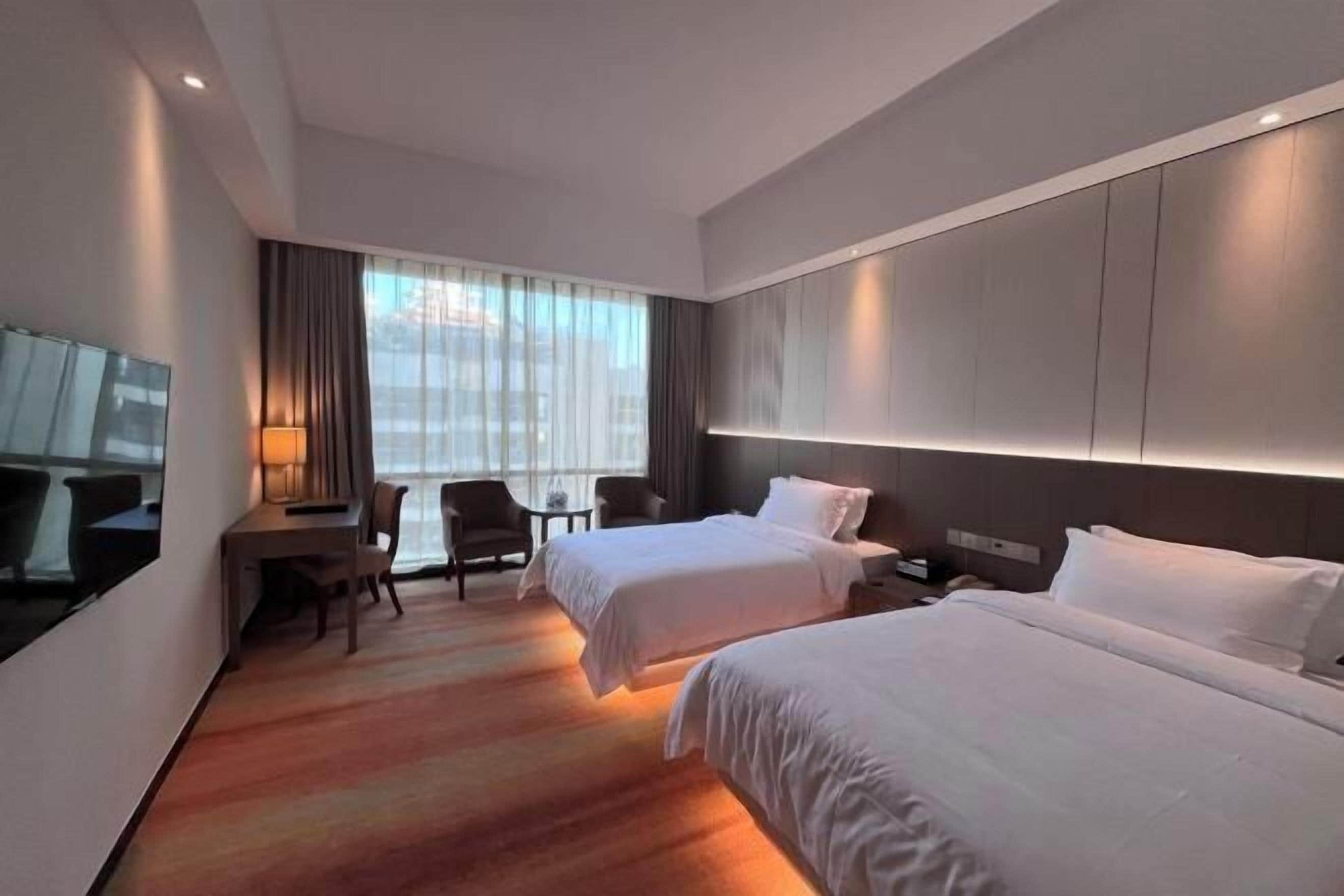 Habitación de hotel con dos camas, un escritorio con silla, televisión y un ventanal con cortinas.