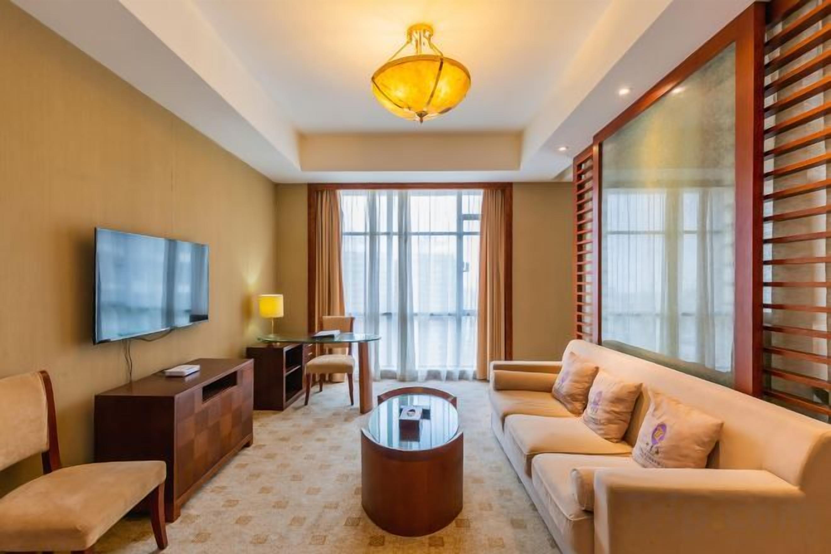A hotel room with a flat-screen TV, a sofa, a coffee table, a desk with a lamp, and a large window with blinds.