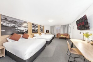 Habitación de hotel con tres camas, un escritorio, un sofá y un televisor montado en la pared.