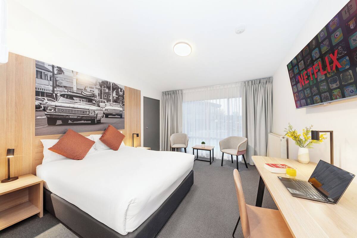 A hotel room with a bed, a desk with a laptop, a chair, a small table with a vase, and a wall with a 'NETFLIX' poster.
