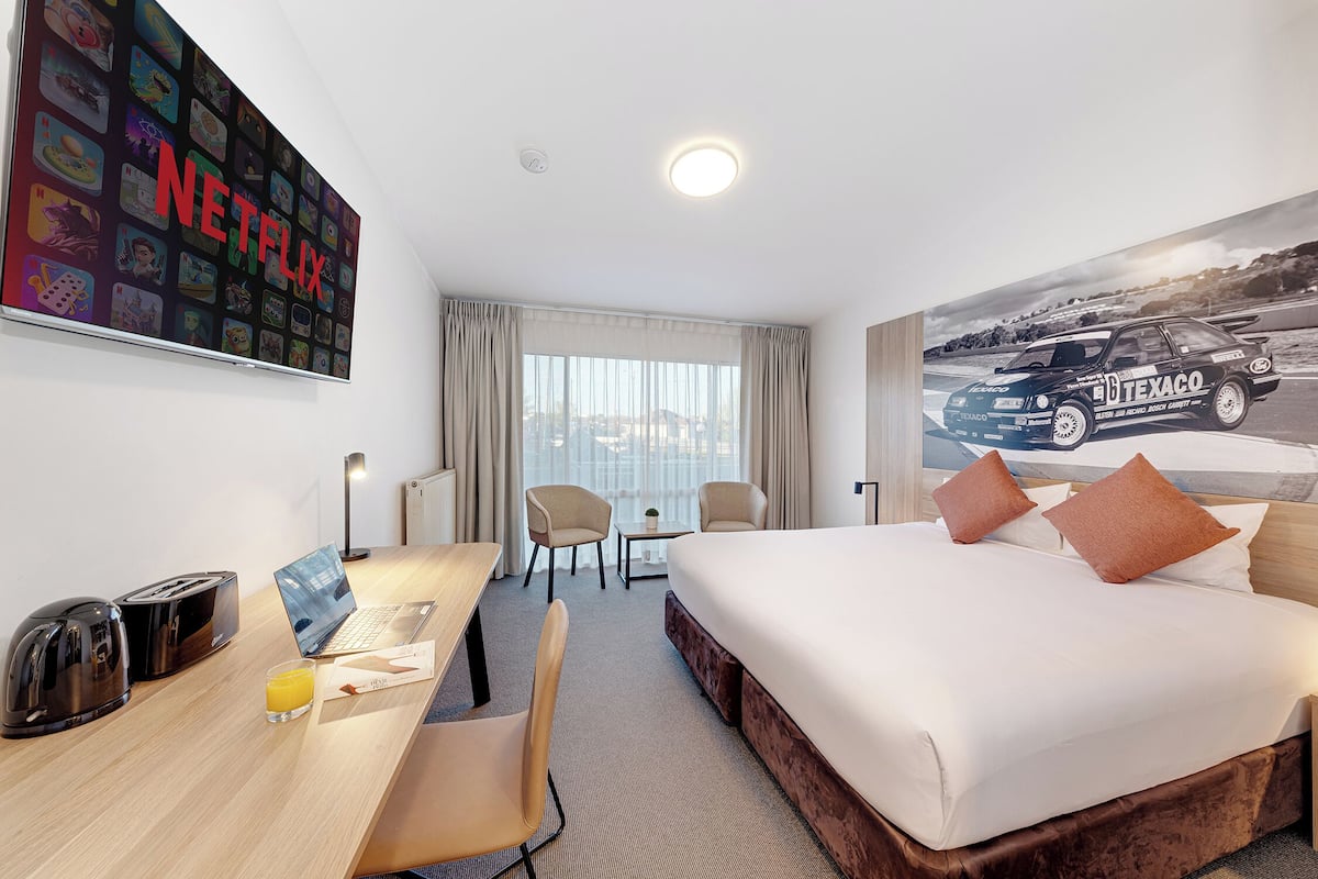 A hotel room with a bed, desk, chairs, a laptop, a toaster, a glass of juice, a large Netflix sign, and a racing car poster.