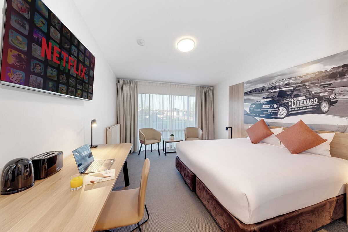 A hotel room with a bed, desk, chairs, a laptop, a toaster, a glass of juice, a large Netflix sign, and a racing car poster.