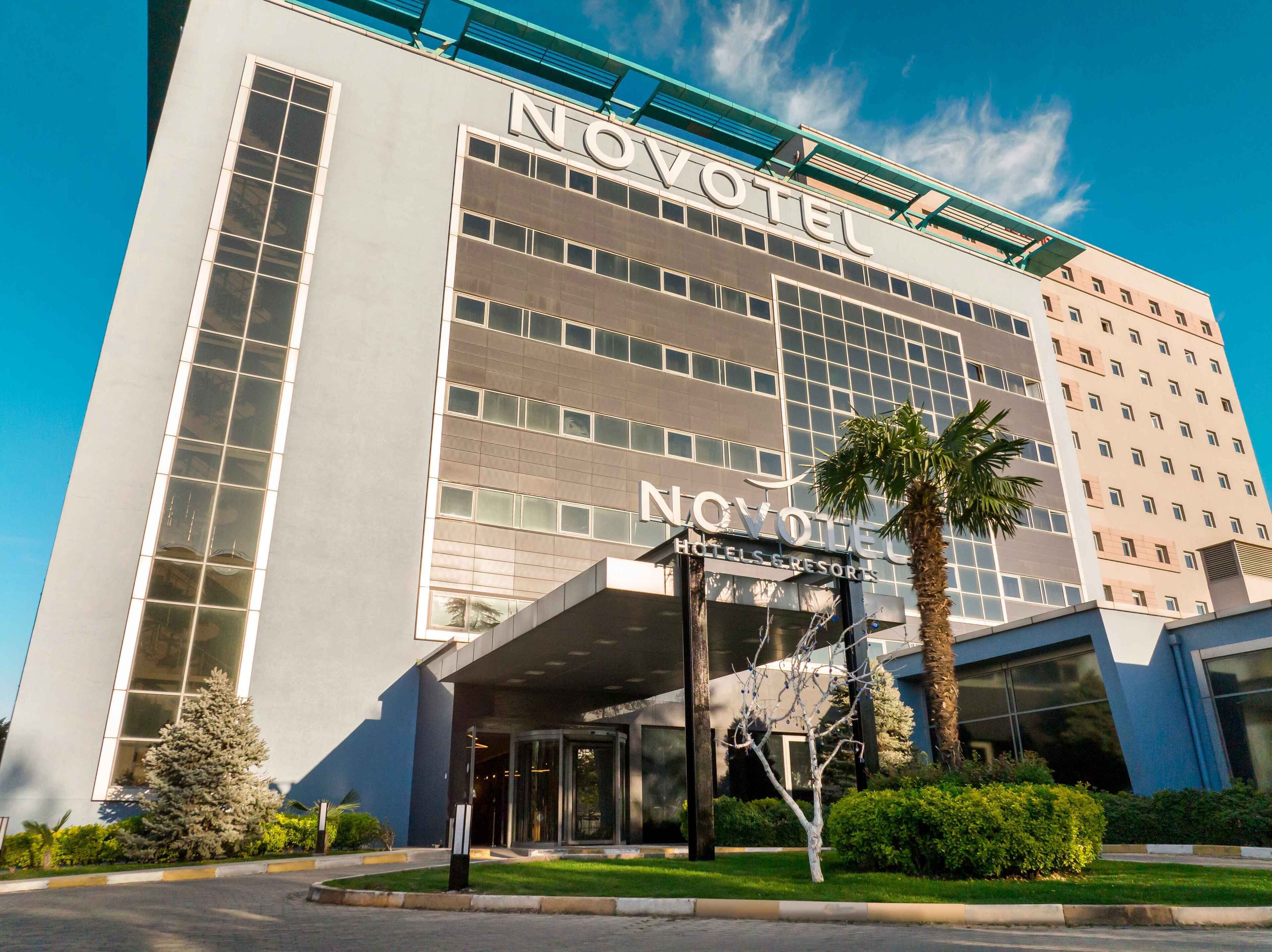 Un moderno edificio de hotel con un llamativo letrero 'NOVOVOTE', grandes ventanales y un área de entrada bien cuidada con una palmera.