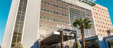 Un moderno edificio de hotel con un llamativo letrero 'NOVOVOTE', grandes ventanales y un área de entrada bien cuidada con una palmera.