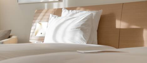 Una cama bien hecha con sábanas y almohadas blancas, cabecera de madera y mesita de noche con una lámpara.