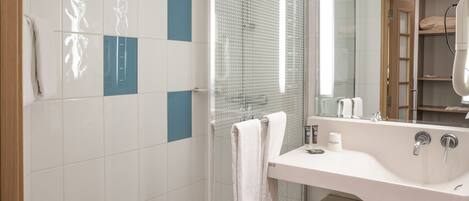 Un baño moderno con cabina de ducha de vidrio, un lavamanos blanco con espejo y un estante de madera con toallas.