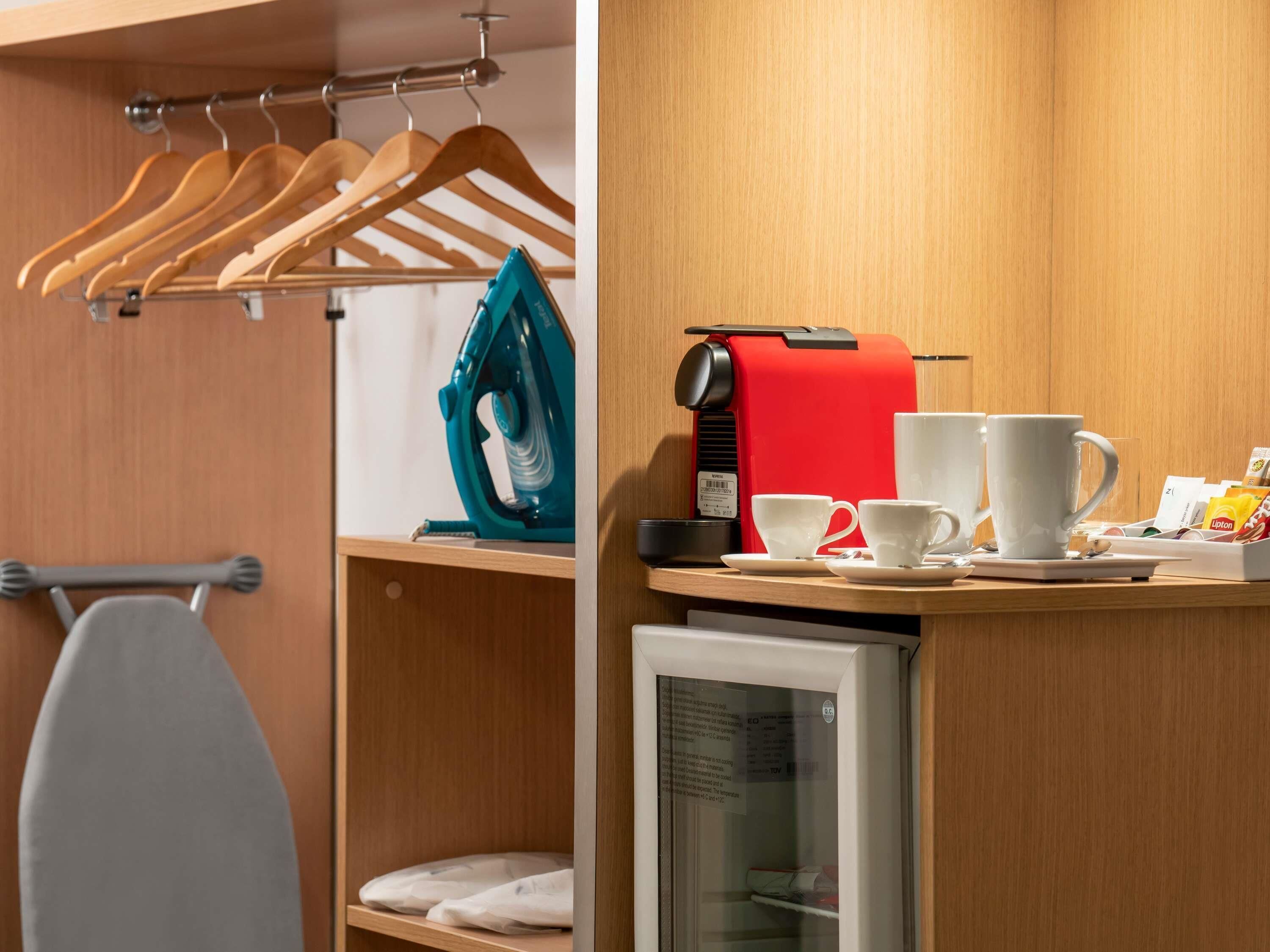 A hotel room with a wooden wardrobe, a small refrigerator, a coffee machine, and a set of white cups and saucers.