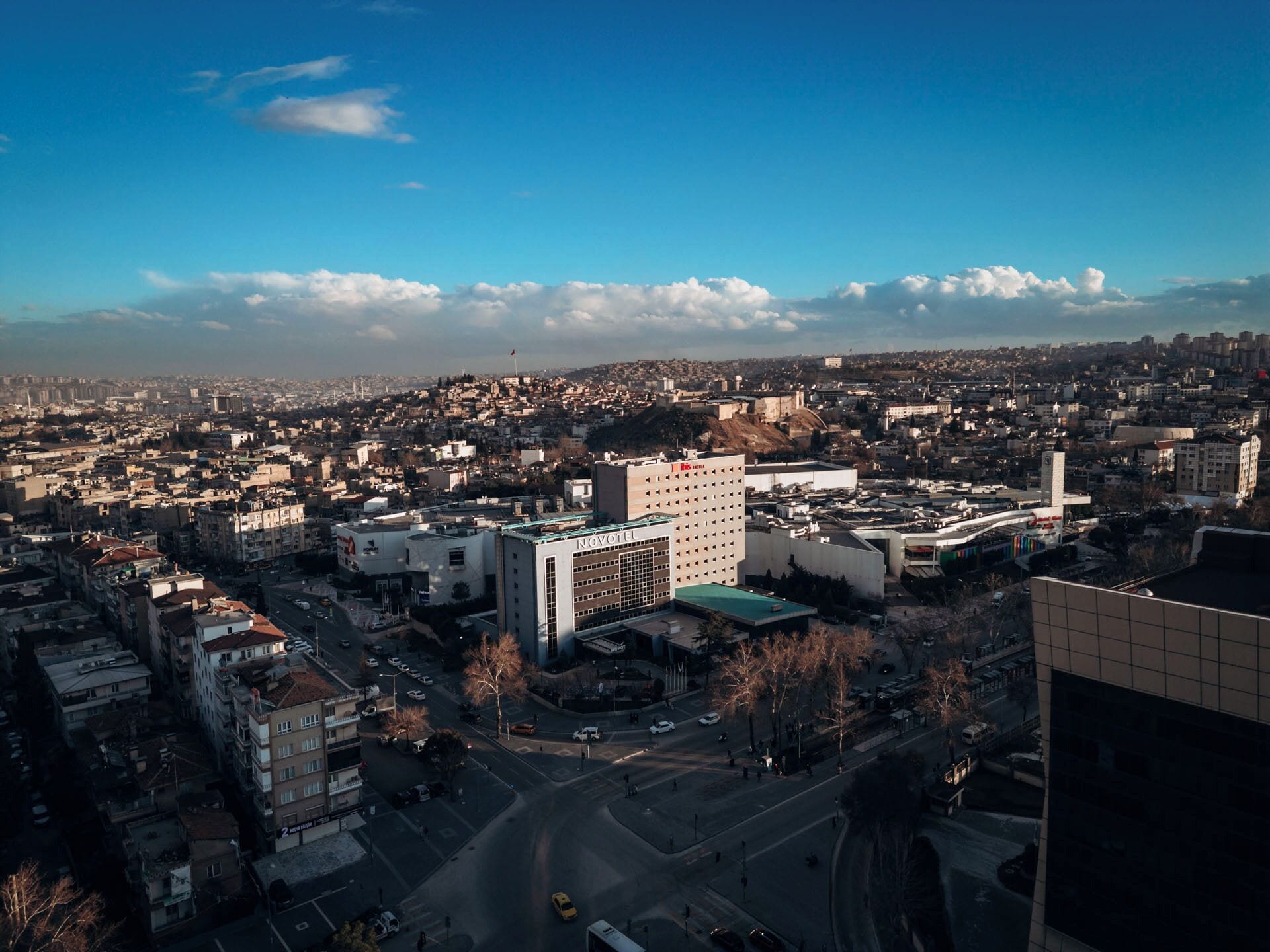 Un paisaje urbano con un hotel destacado, identificado como 'NOVOTEL', en el centro, rodeado de otros edificios y un cielo despejado.