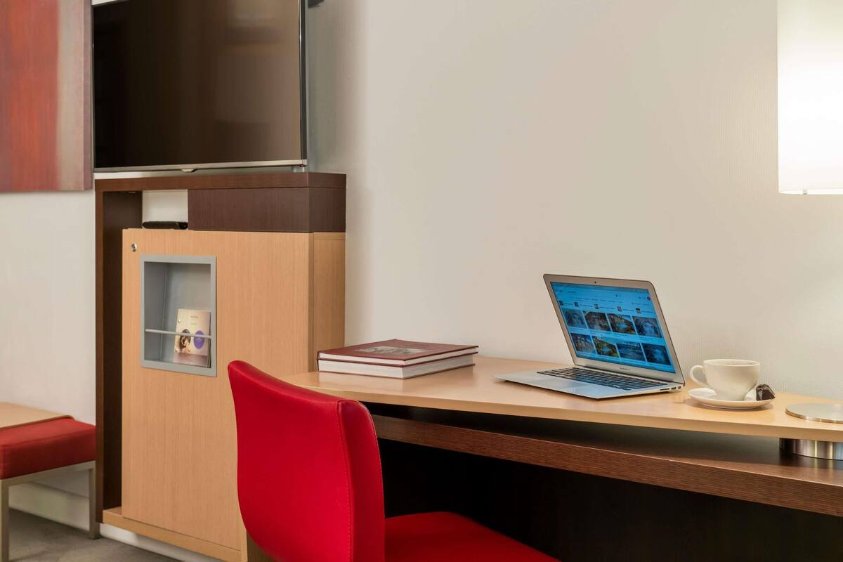 A hotel room with a desk, a laptop, a cup, and a TV.