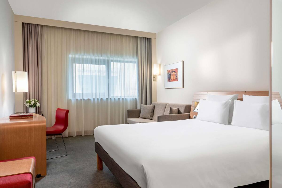 A hotel room with a large bed, a desk, a chair, a sofa, and a window with curtains.
