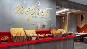 Mostrador de panadería con pan y pasteles, pared con la palabra 'nefas' y 'KESIF MUTFAGI' escritas, y un diseño de interiores moderno con elementos de madera y una encimera roja.