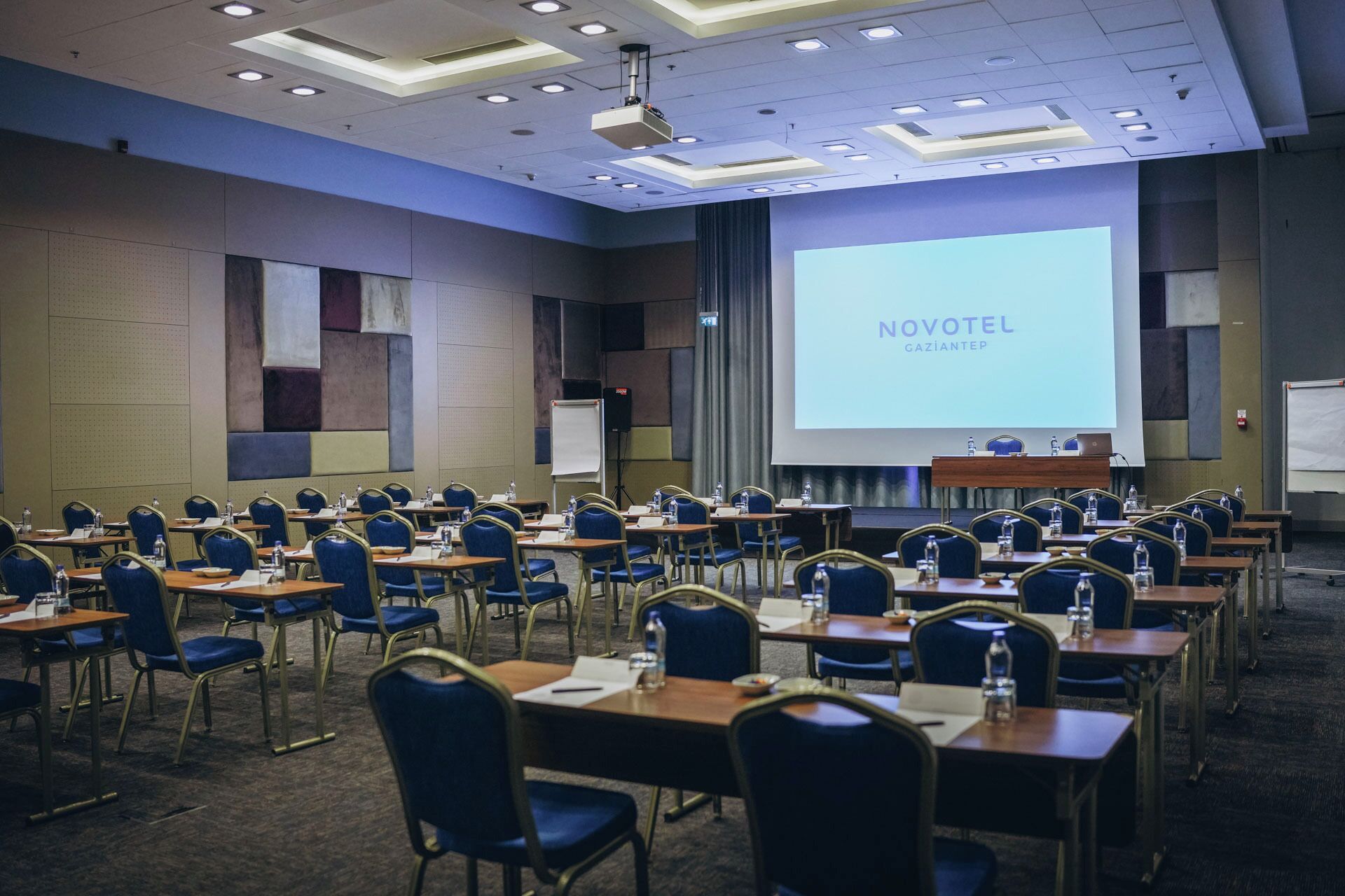 Un salón de conferencias con filas de mesas y sillas, una pantalla de proyector mostrando 'NOVOTEL GAZIANTEP' y una pizarra.