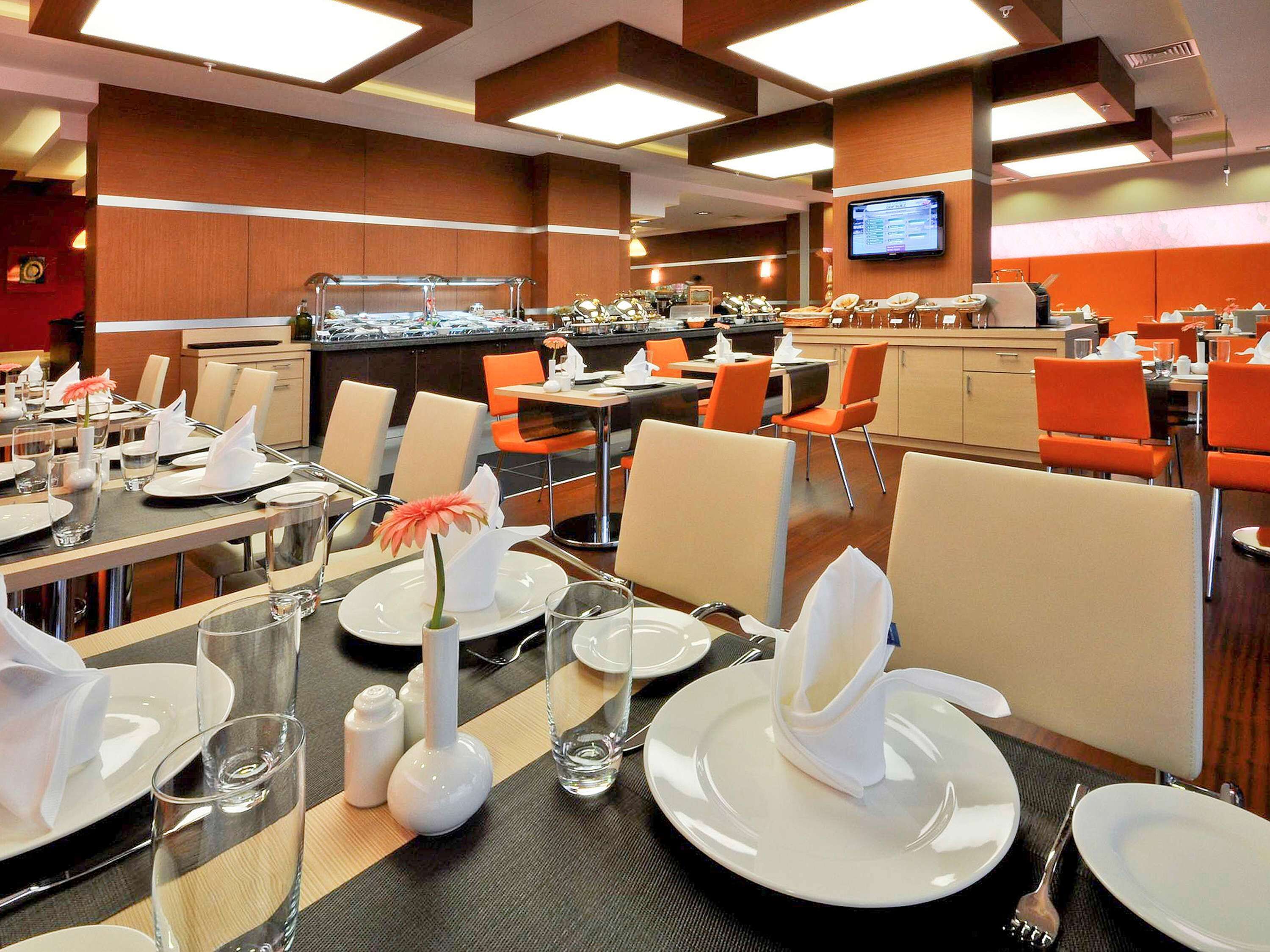 Zona de comedor de hotel con mesas dispuestas para los huéspedes, una barra de buffet al fondo y un televisor montado en la pared.
