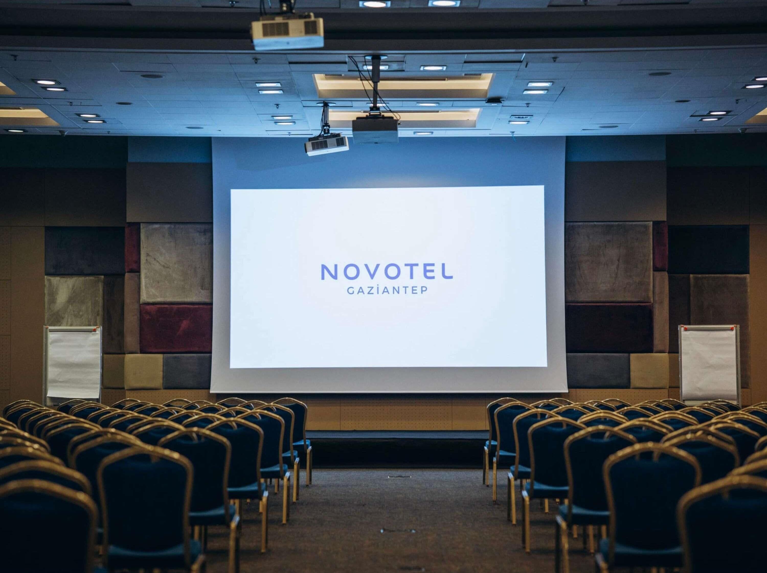 Ein Konferenzraum mit Reihen von Stühlen, einem großen Bildschirm, der 'NOVOTEL GAZIANTEP' zeigt, und einem an der Decke befestigten Projektor.