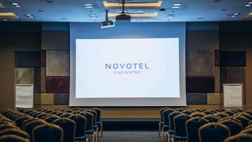 Ein Konferenzraum mit Reihen von Stühlen, einem großen Bildschirm, der 'NOVOTEL GAZIANTEP' zeigt, und einem an der Decke befestigten Projektor.