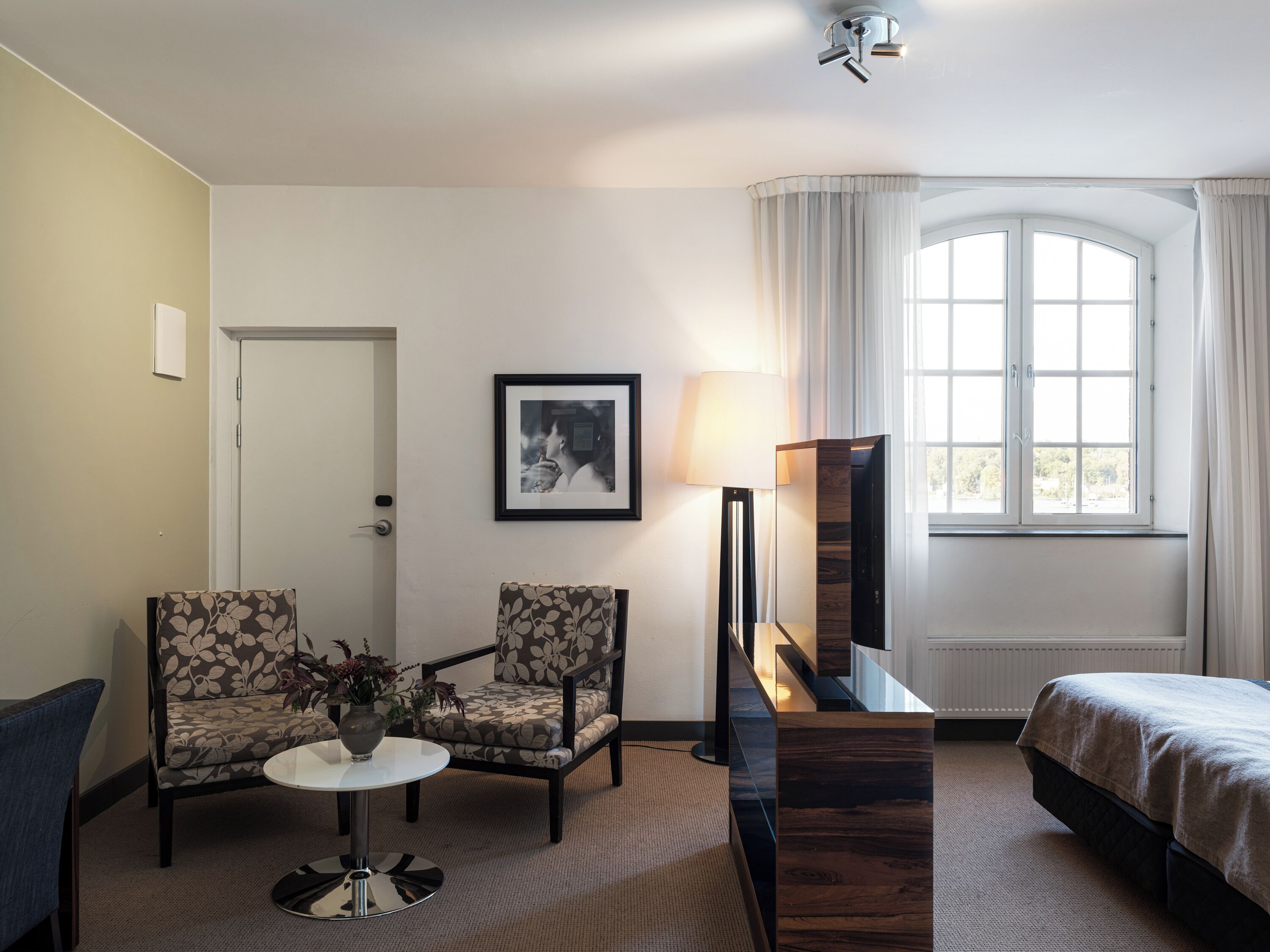A hotel room with two armchairs, a small round table with a vase, a bed, a large mirror, and a window with curtains.