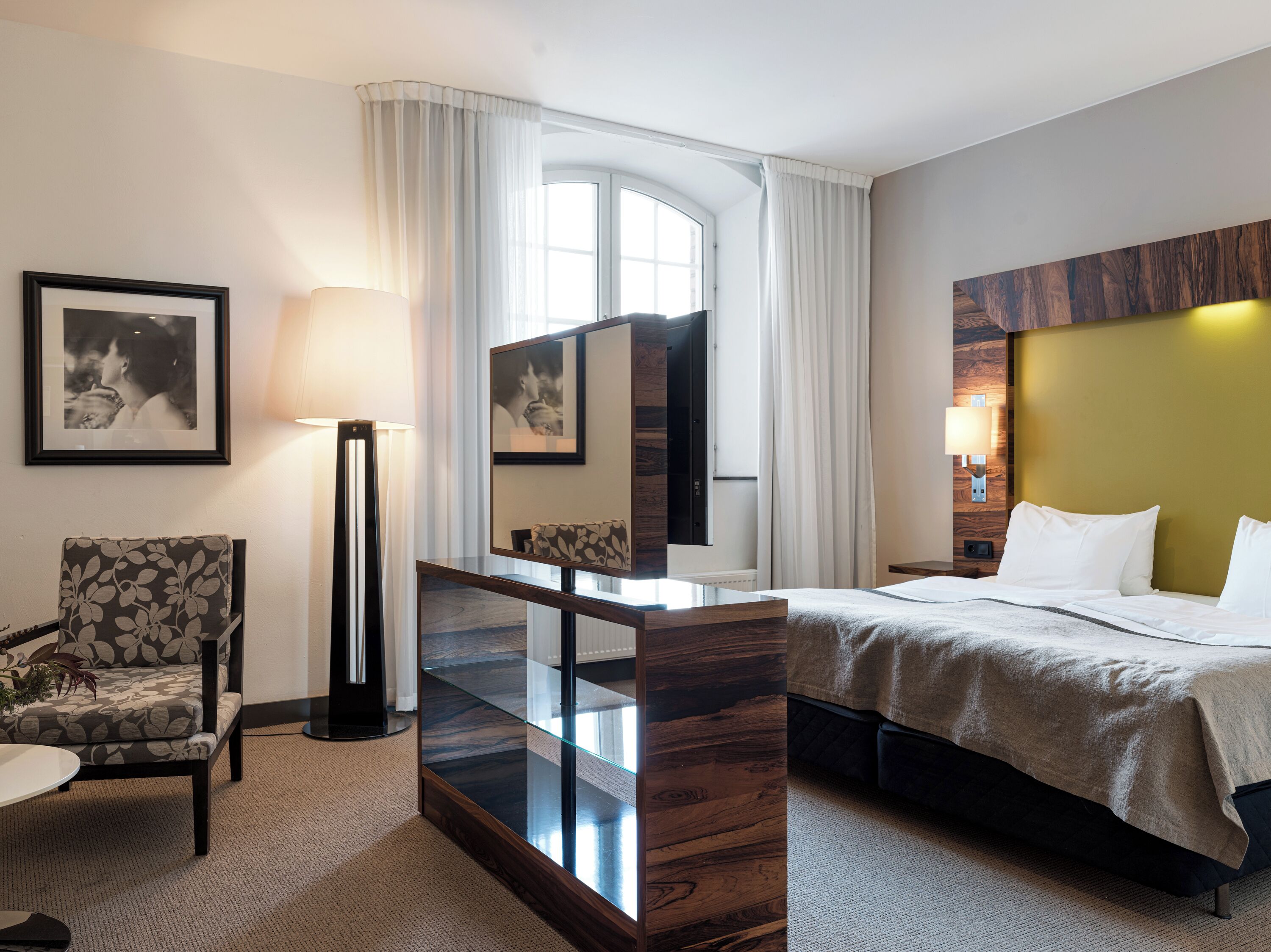 A hotel room with a bed, a chair, a desk with a lamp, a large mirror, and a window with curtains.