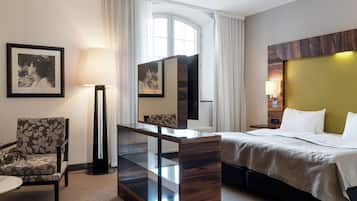 A hotel room with a bed, a chair, a desk with a lamp, a large mirror, and a window with curtains.