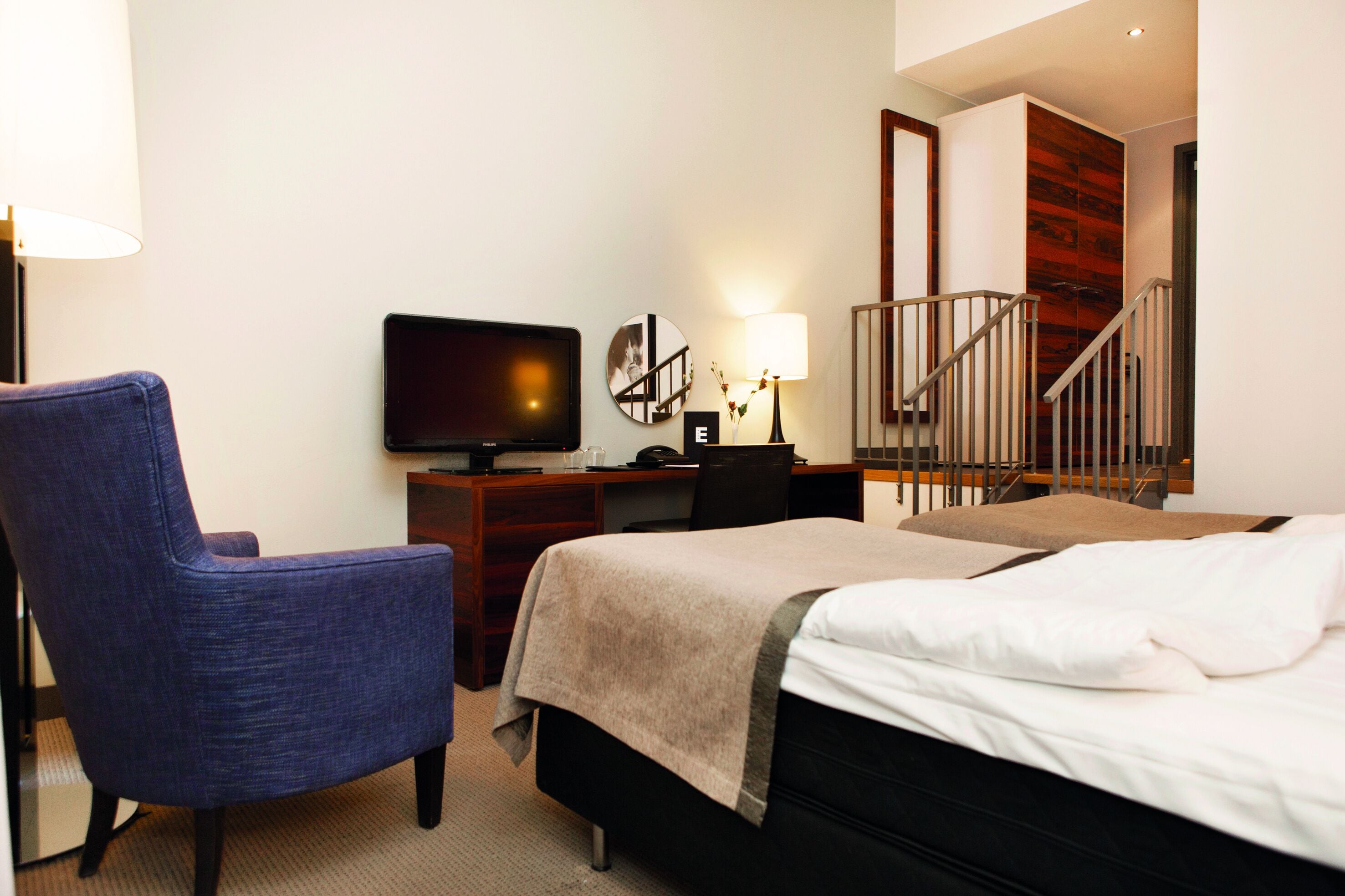 A hotel room with a bed, a desk with a television, a chair, a lamp, and a staircase.