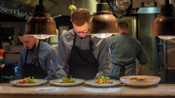 Un chef prepara comida en una cocina profesional con iluminación superior y electrodomésticos de acero inoxidable.