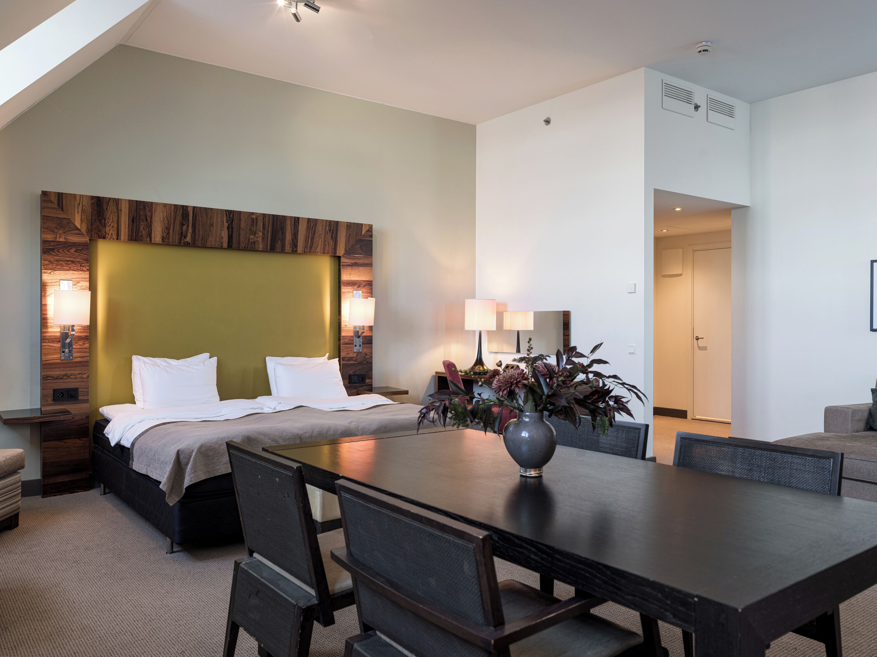 Ein Hotelzimmer mit Bett, Esstisch, Stühlen, einer Vase mit Blumen und einer Lampe.