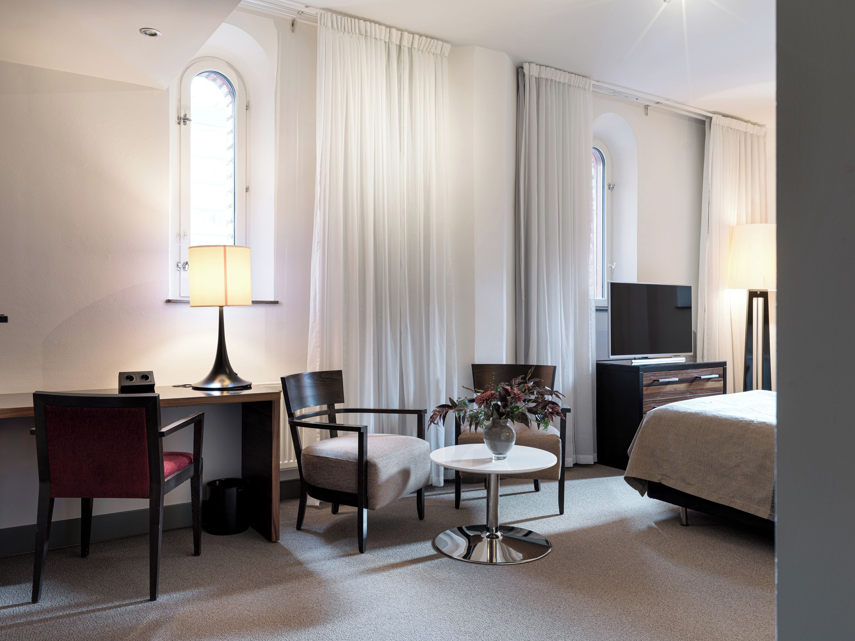 Et hotelværelse med en seng, et skrivebord med lampe, to stole, et lille bord med en vase og et fjernsyn.