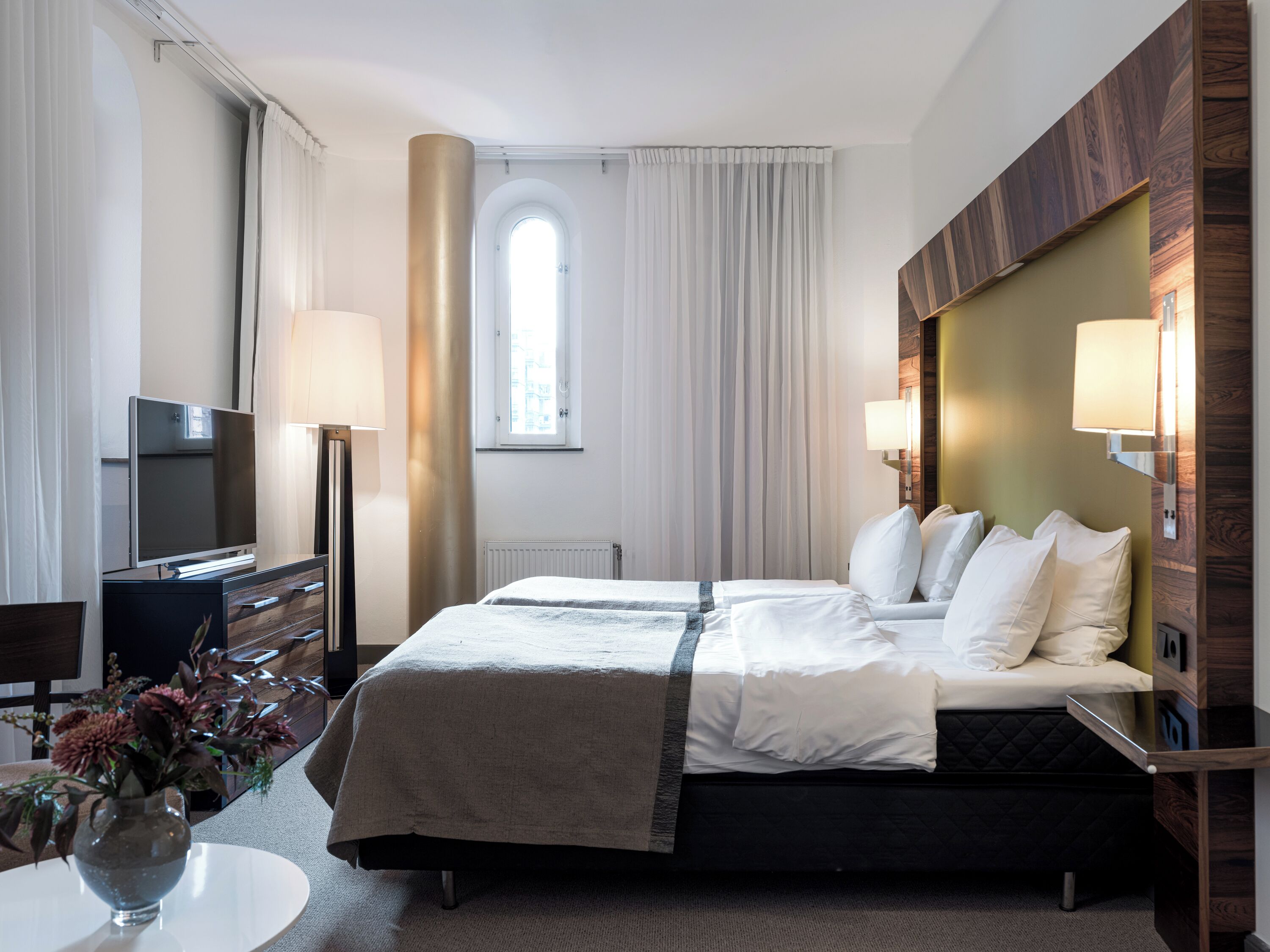 Et hotelværelse med seng, fjernsyn, skrivebord og et vindue med gardiner.
