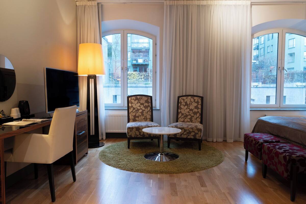 Habitación de hotel con escritorio, dos sillones, una mesa redonda, cama y ventanas grandes con cortinas.