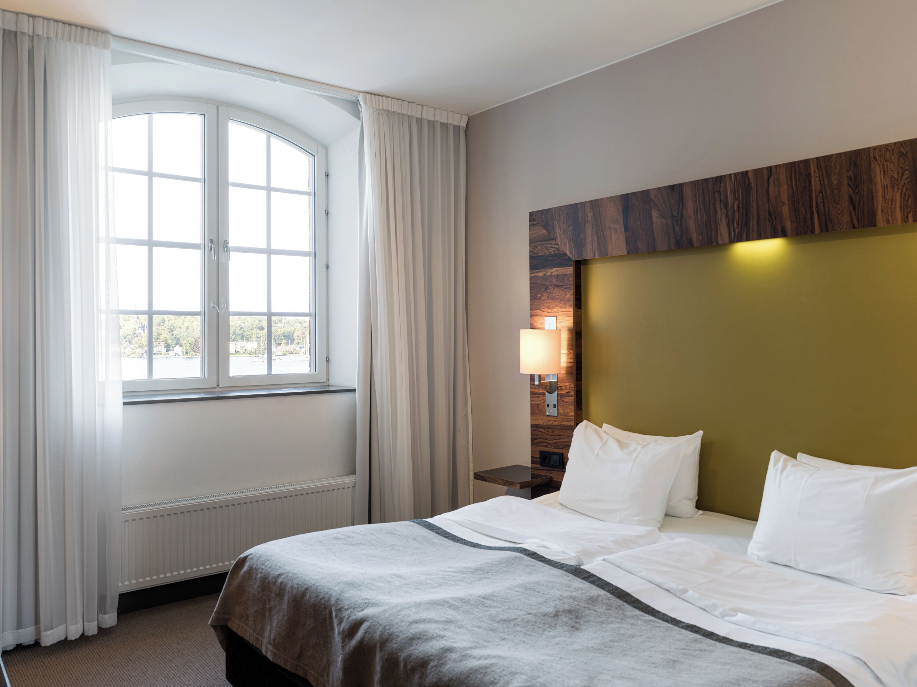 Habitación de hotel con un ventanal grande, una cama con ropa blanca, una mesita de noche con lámpara y un cabecero de madera.
