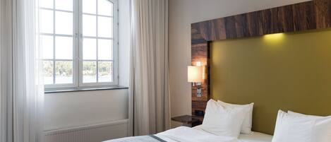 A hotel room with a large window, a bed with white linens, a nightstand with a lamp, and a wooden headboard.