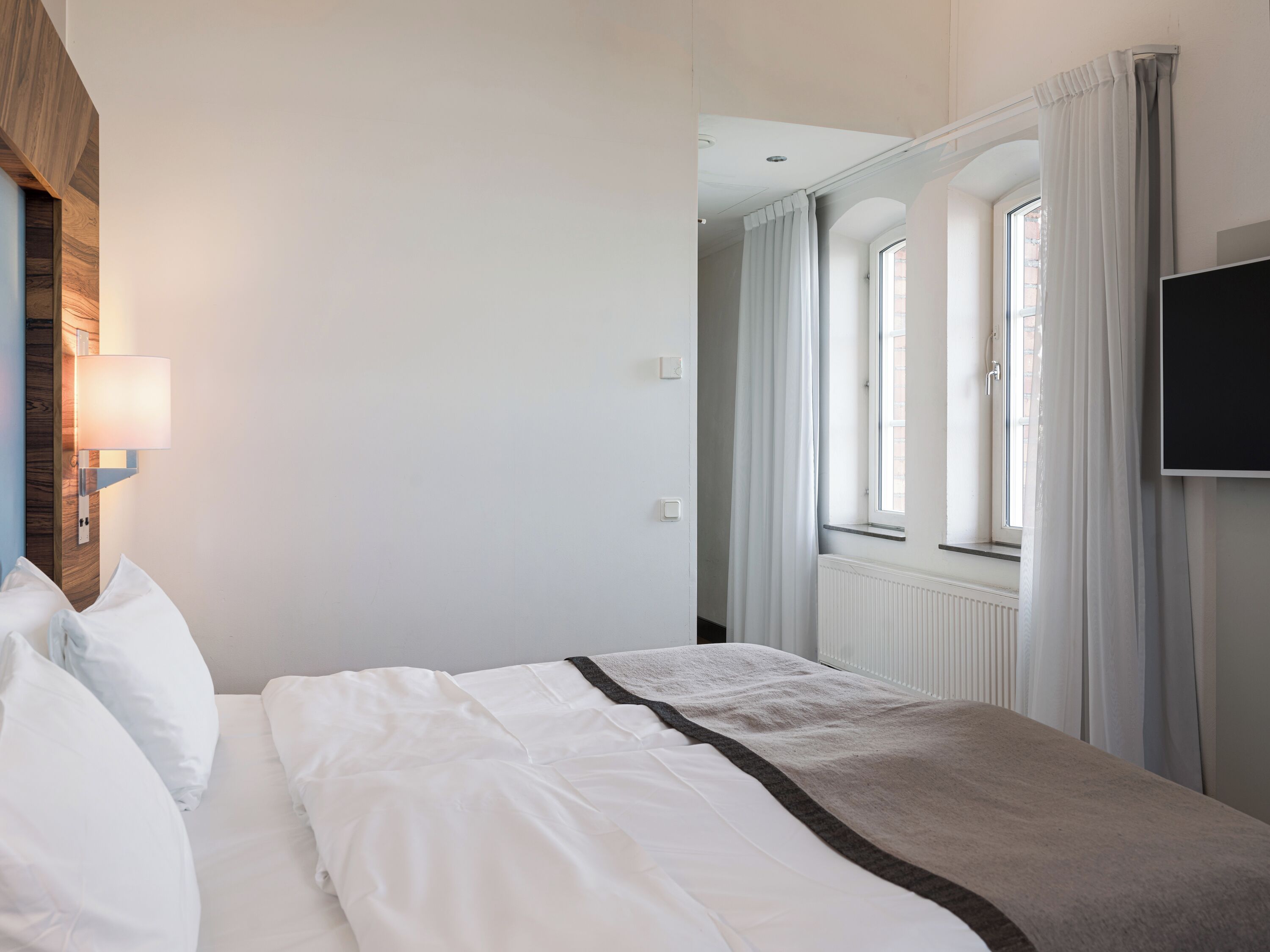 A hotel room with a bed, a wall-mounted TV, and a window with curtains.