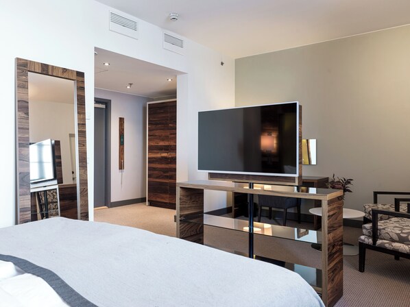 Una habitación de hotel moderna con una cama grande, un televisor de pantalla plana sobre un soporte de madera, una zona de estar con una mesa y sillas, y un espejo en la pared.