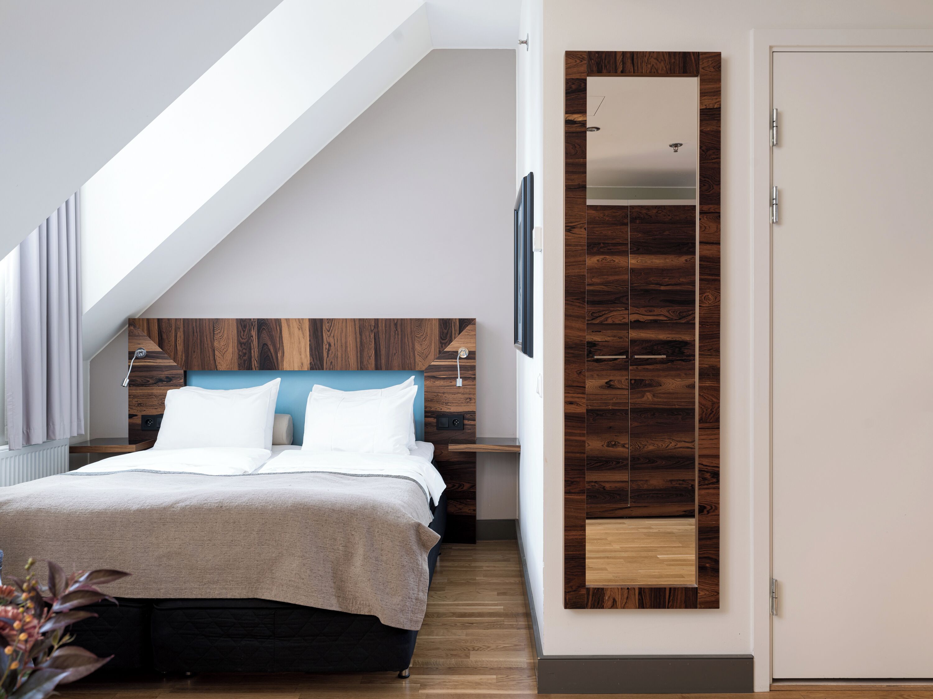 A modern bedroom with a sloped ceiling, a wooden headboard, a bed with white linens, a bedside table, and a mirror on the wall.