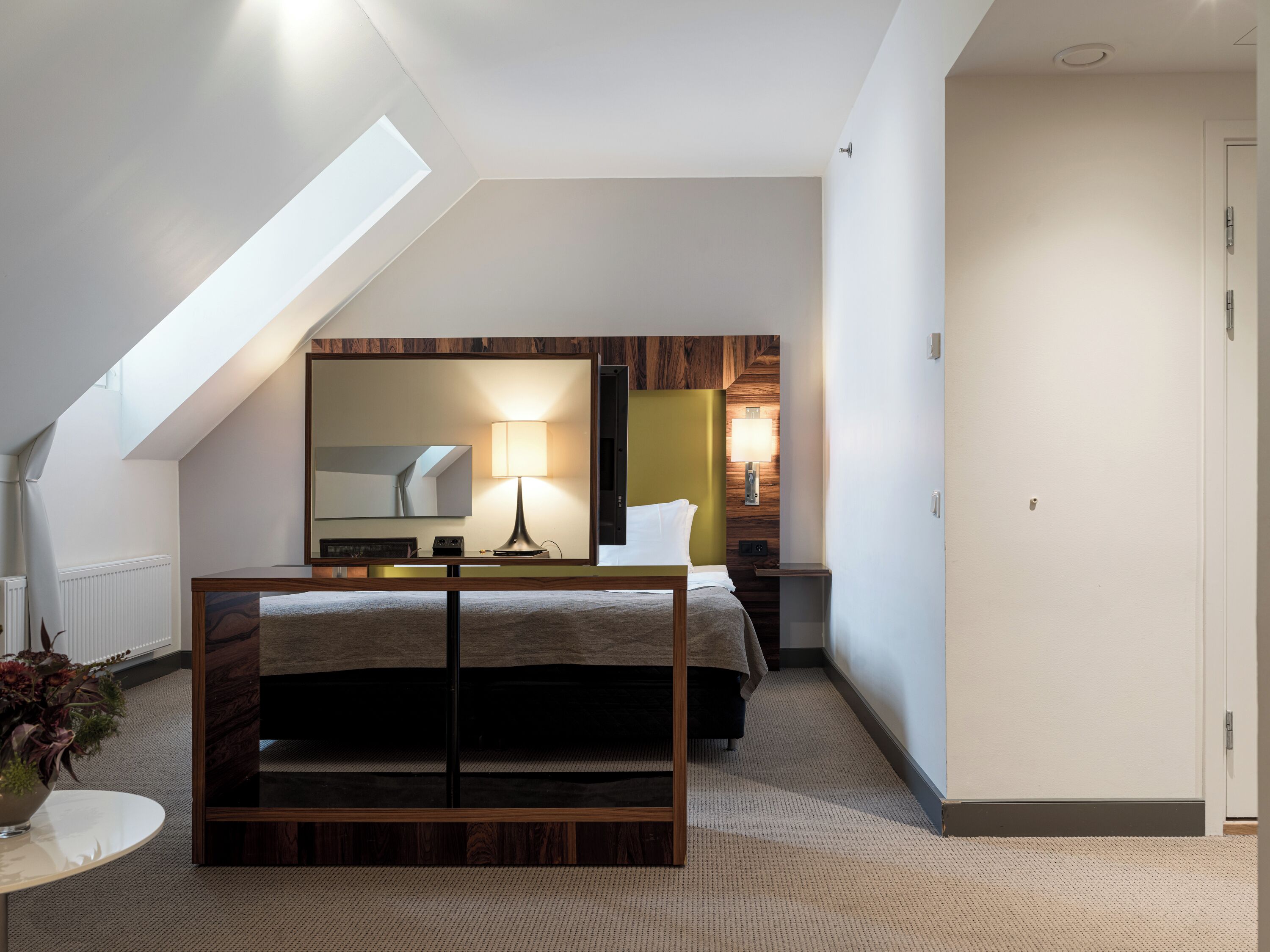 A modern hotel room with a sloped ceiling, a bed with a wooden frame, a bedside table with a lamp, a mirror, and a view of the hallway.